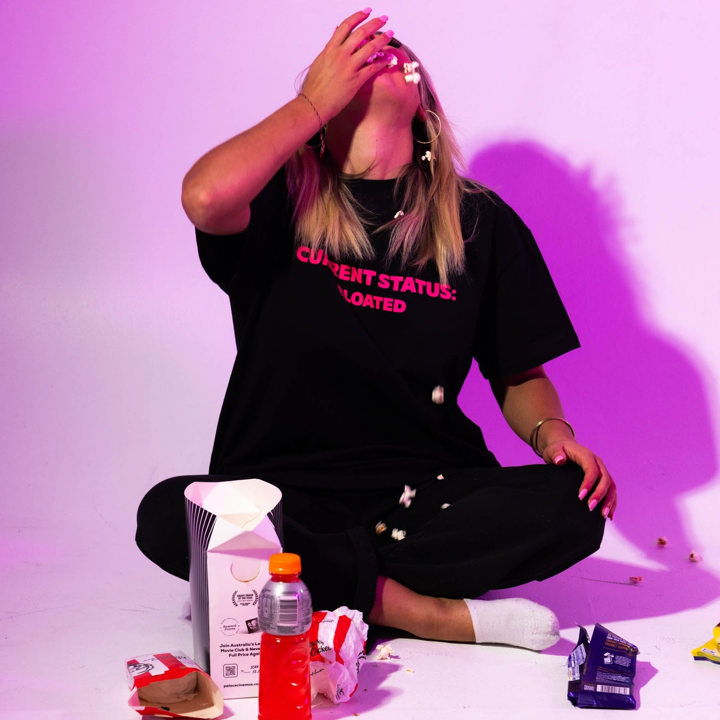 Person wearing a black t-shirt with text, sitting on the floor with various items around, against a purple background.