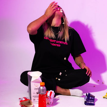 Person wearing a black t-shirt with text, sitting on the floor with various items around, against a purple background.