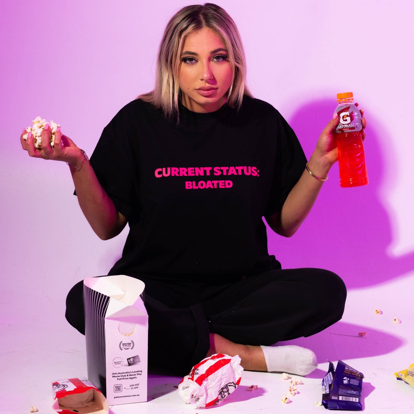 Person wearing a black t-shirt with 'CURRENT STATUS: BLOATED' text, sitting on the floor with snacks and drinks against a pink background.