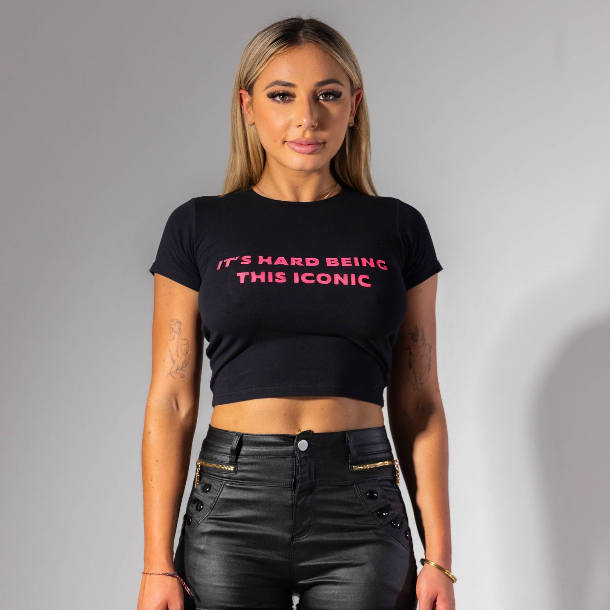 Woman wearing a black crop top with pink text and black leather shorts on a gray background