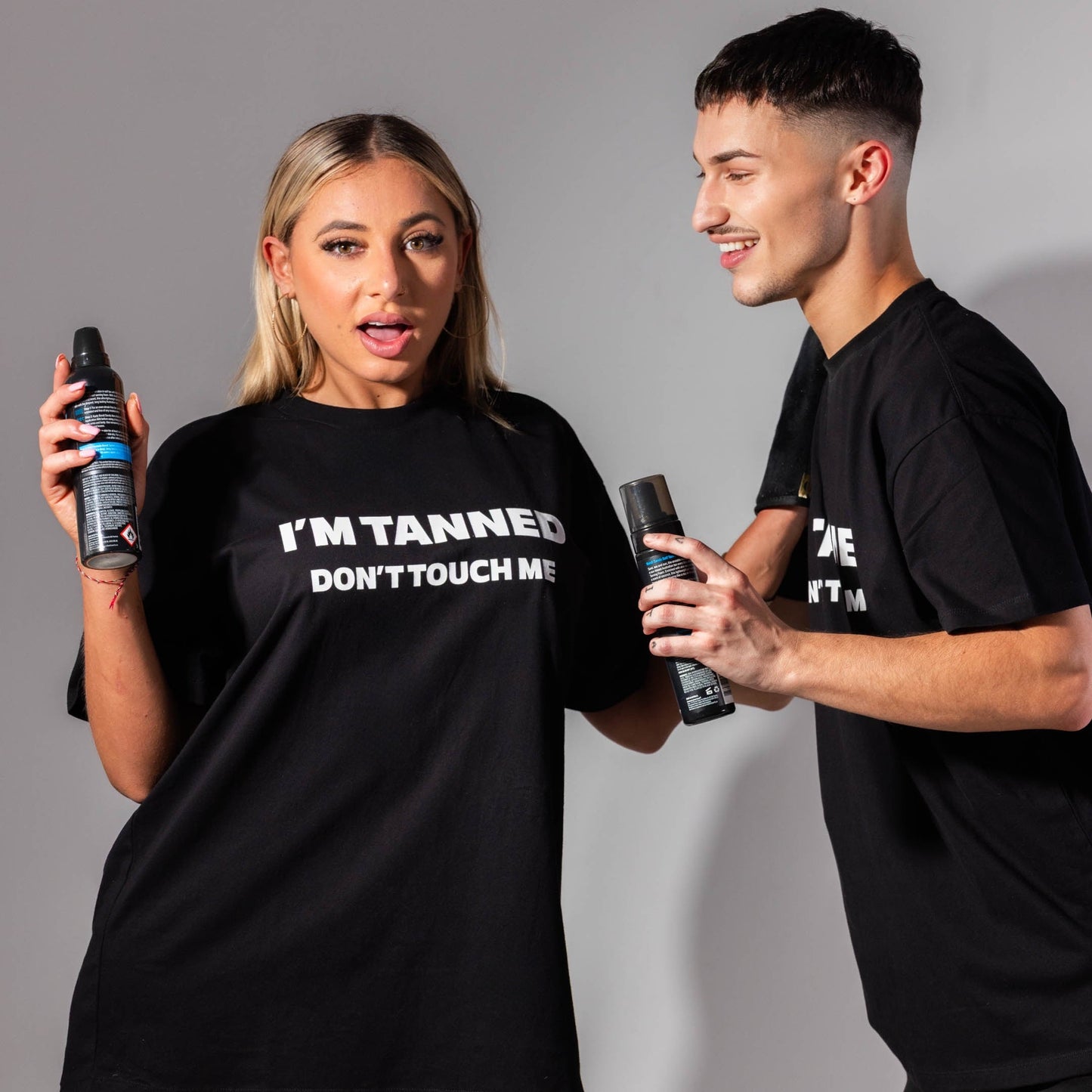 Two people wearing black t-shirts with humorous text, holding tanning products against a gray background.